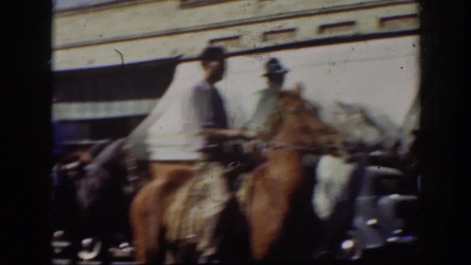 Two blurred horseback riders in hats move past storefronts and automobiles in this 1939 California 8mm home-movie frame.