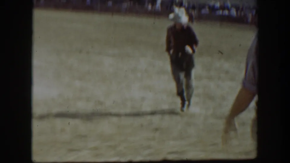 A cowboy in a light hat and dark clothing rides away across a California rodeo arena with spectators in the distance.