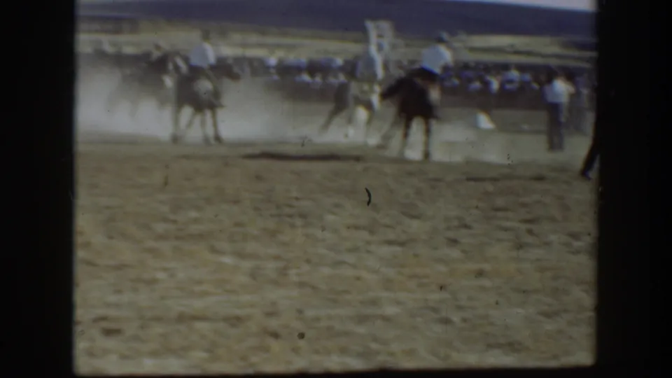 A distant rodeo moment shows horses, riders, dust, and spectators around the arena as the action continues.