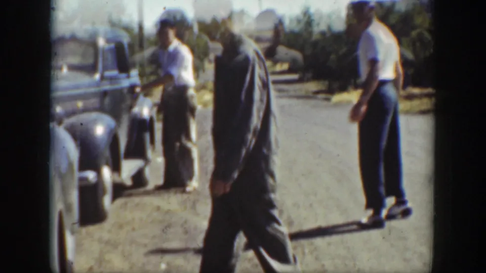 1939 California roadside frame showing men in casual work clothes near older automobiles on a dusty town street.