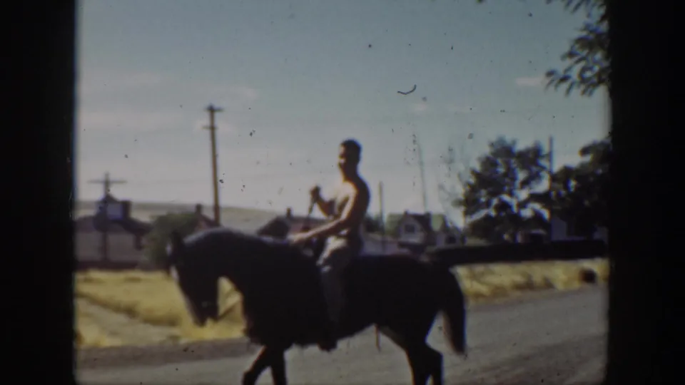 1939 California home-movie frame showing a man riding a dark horse along a town road with utility poles and trees in the background.
