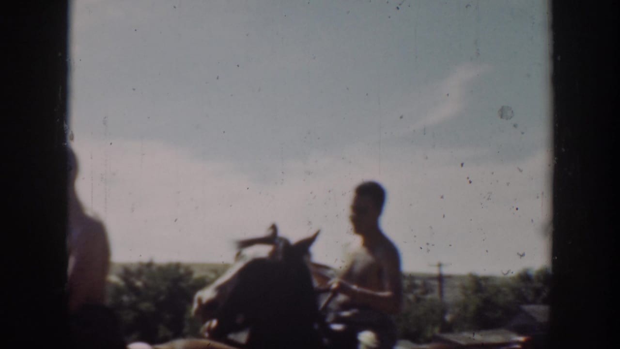 1939 California home-movie frame showing a shirtless man on horseback passing a dark doorway or window edge near open fields.