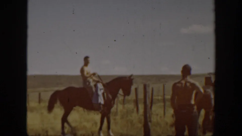 A rider sits on a horse in an open field while another shirtless man stands nearby, with fence posts and rural land behind them.
