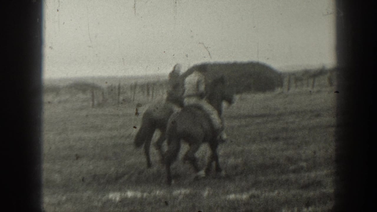 In 1939 Montana home-movie footage, two riders on horseback move through an open field with a fence line and haystack behind them.