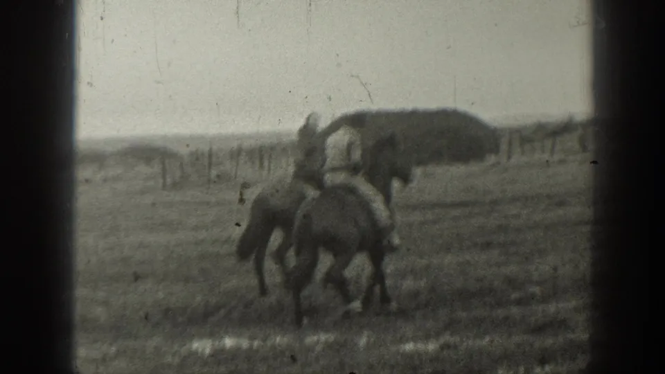 In 1939 Montana home-movie footage, two riders on horseback move through an open field with a fence line and haystack behind them.