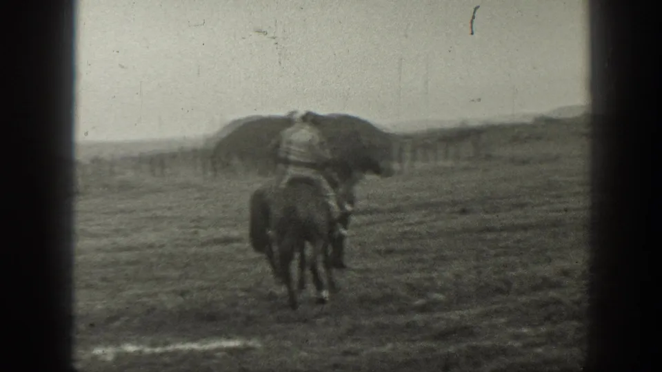 A mounted rider appears in a Montana field, facing away from the camera with a large haystack and fence line in the background.