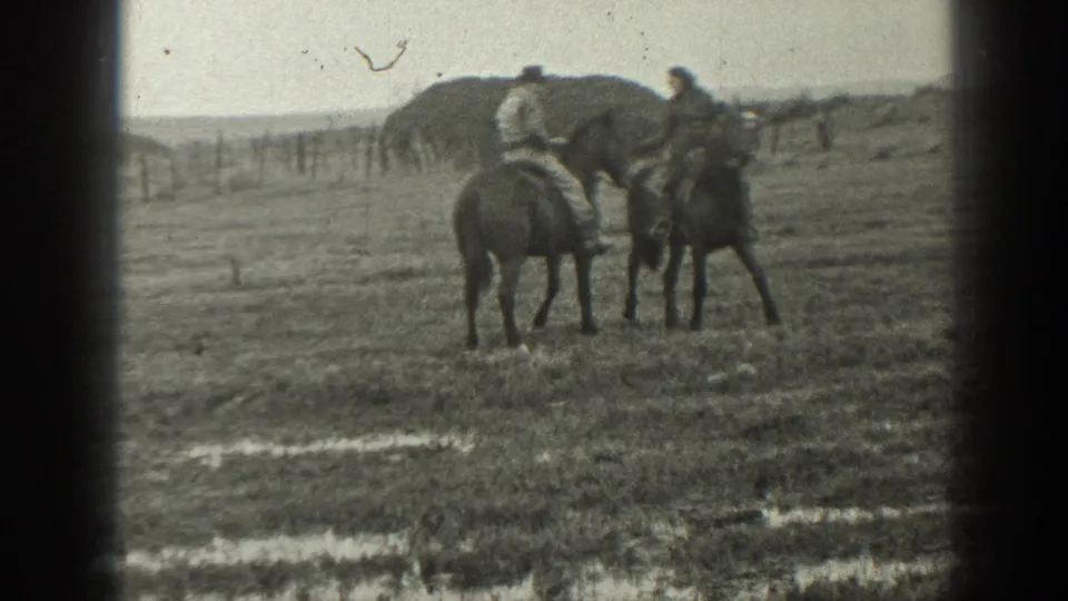 Two adults on horseback appear to pause or converse in a rural Montana field with haystacks and fencing behind them.