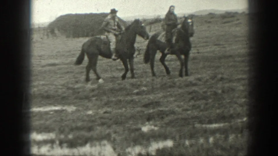 Two mounted riders in 1939 Montana stand in an open field, one wearing a hat, with a haystack and low hills visible behind them.