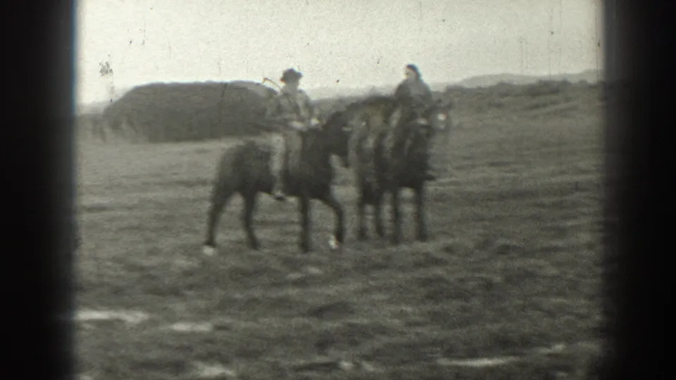 Two adults ride horses in a Montana ranch landscape, with soft 8mm edges and a low hill or mound in the background.