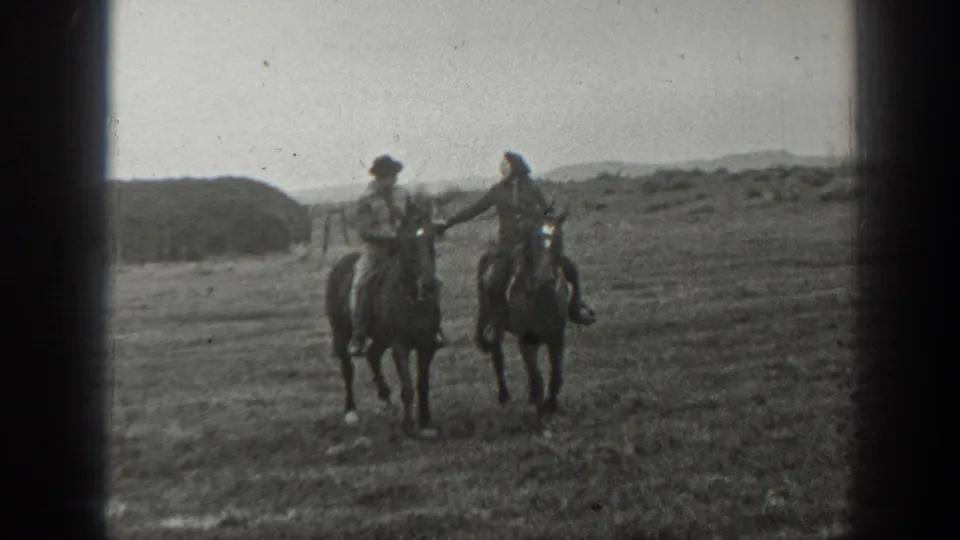 Two adults on horseback ride toward the camera in a rural Montana setting, with one rider reaching toward the other.