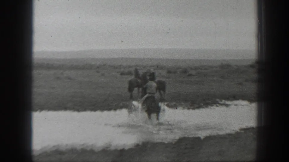 Distant 1939 Montana 8mm frame showing a horseback rider guiding horses across shallow water in open terrain.