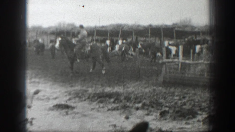 Soft black-and-white 8mm frame of a rider moving near cattle, puddled ground, and ranch fencing in Montana.