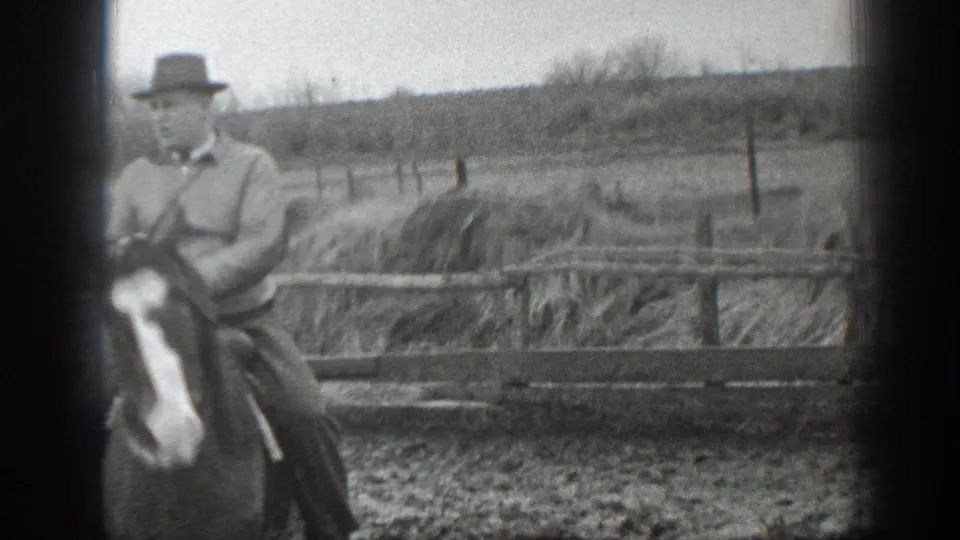 A man in period farm clothing rides a horse near wooden fencing in the Montana farmyard, with pasture and brush beyond.