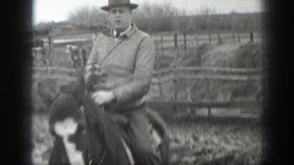 Close 1939 Montana home-movie frame of a man on horseback near wooden pens, muddy ground, and open farm fields.