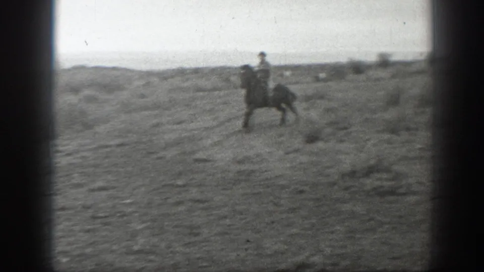 A cowboy on horseback moves across rough Montana prairie in grainy black-and-white 8mm home-movie footage.
