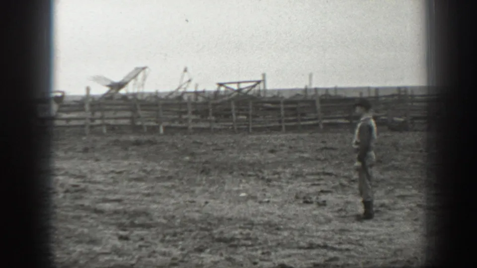 A man stands in a wide 1939 Montana ranch field facing wooden fencing and distant ranch equipment or structures.