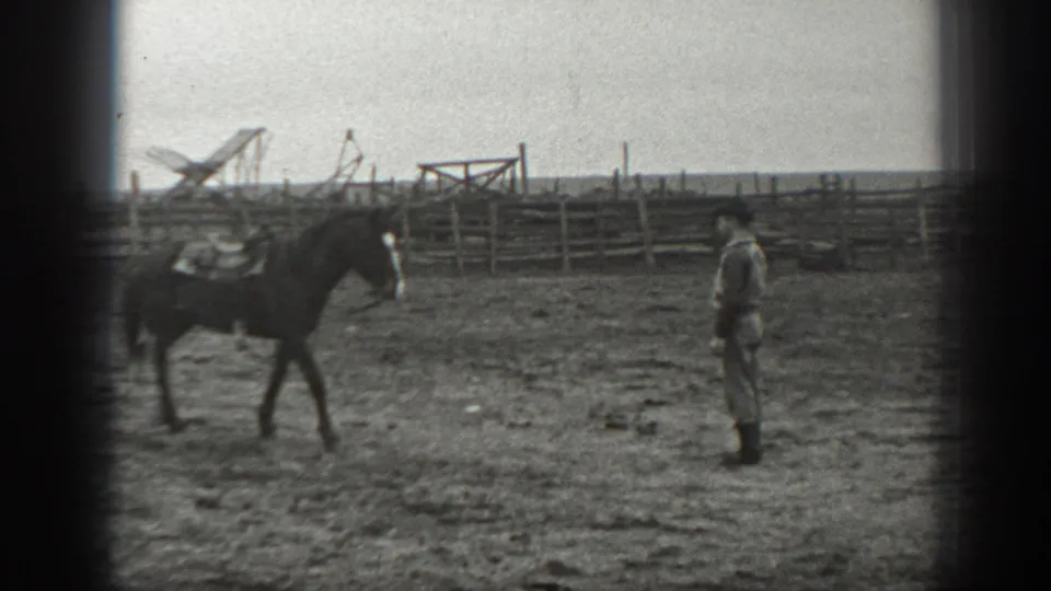 A saddled horse crosses the corral toward a man in period attire, with wooden fencing behind them.