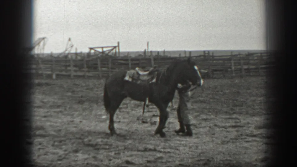 In 1939 Montana ranch footage, a man in work clothes stands close to a saddled dark horse near rail fencing.