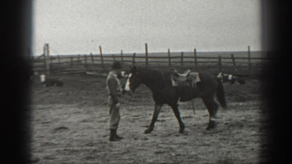 A man in light ranch clothing stands beside a saddled horse, with fencing and distant livestock visible behind them in Montana footage.