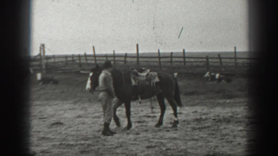 In 1939 Montana home-movie footage, a man leads a saddled horse through a dusty fenced yard with open land behind.