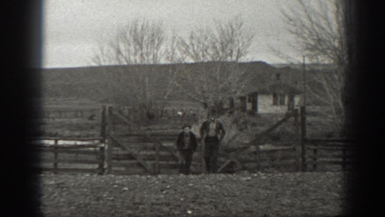 Two men in hats stand by a wooden farm gate in rural Montana, with a small outbuilding, trees, and low hills beyond.