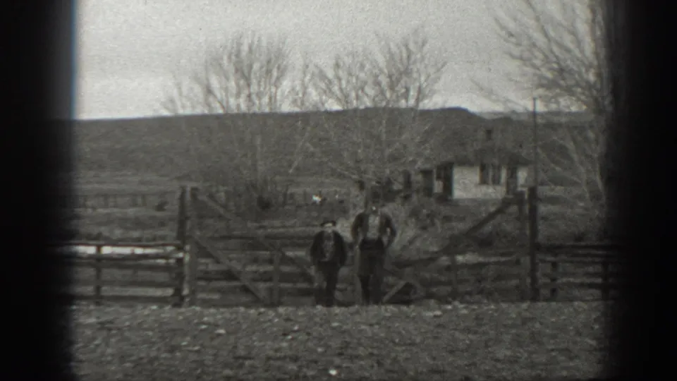 Two men in hats stand by a wooden farm gate in rural Montana, with a small outbuilding, trees, and low hills beyond.