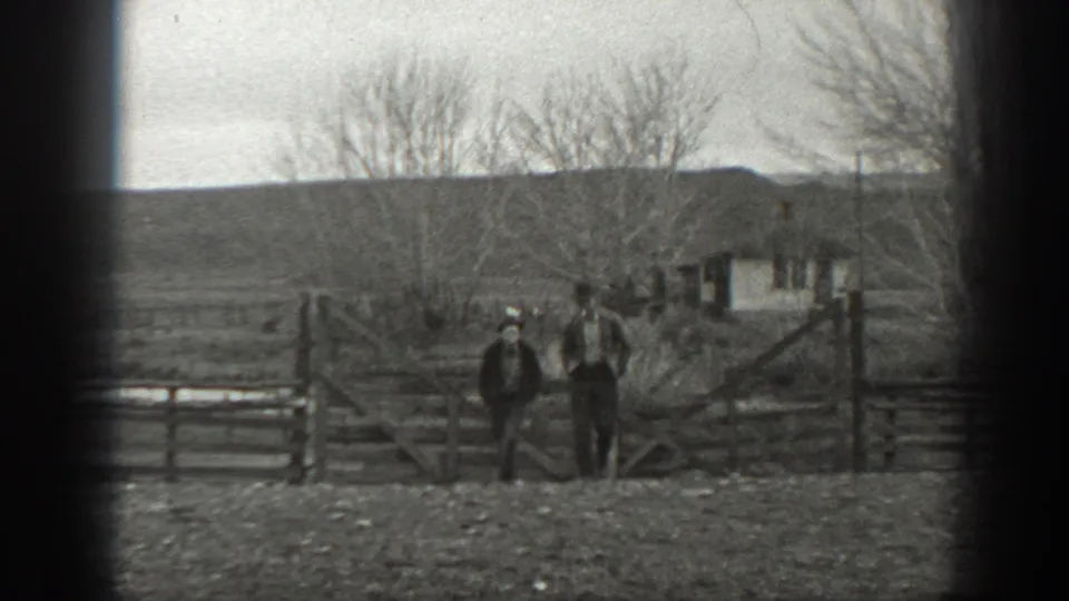 The home-movie frame shows two men standing at a wooden gate on a Montana farm, with bare trees and a modest outbuilding behind them.