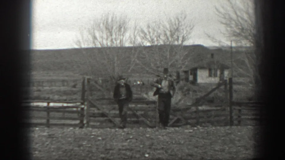 Two men move away from a wooden gate in 1939 Montana footage, framed by fencing, leafless trees, a small outbuilding, and open land.