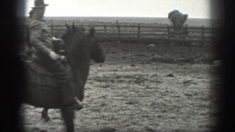 1939 Montana 8mm frame of a mounted man in period clothing riding near a wooden corral fence, with cattle in the distance.