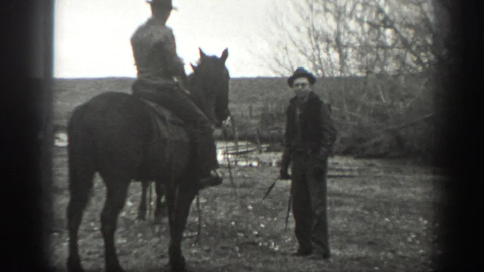 A mounted rider and a standing man in work clothes appear on a Montana ranch, with fencing and bare trees behind them.