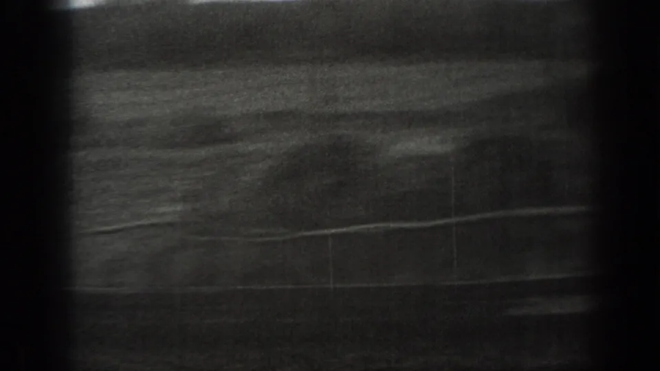 Black-and-white 8mm frame of an open Montana field with a low fence line and dark rural background.
