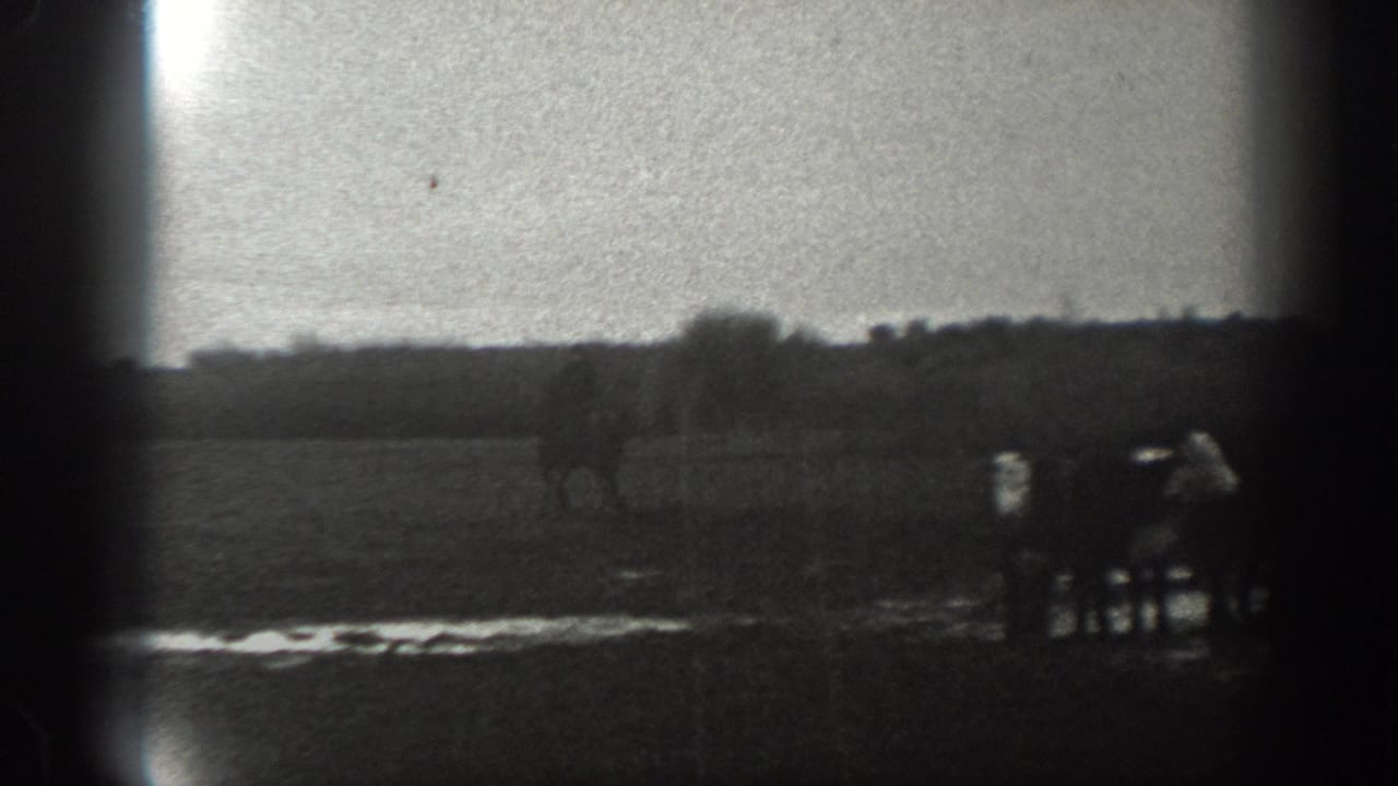 In 1939 Montana home-movie footage, a distant man on horseback moves across a wet rural field beneath a pale sky.
