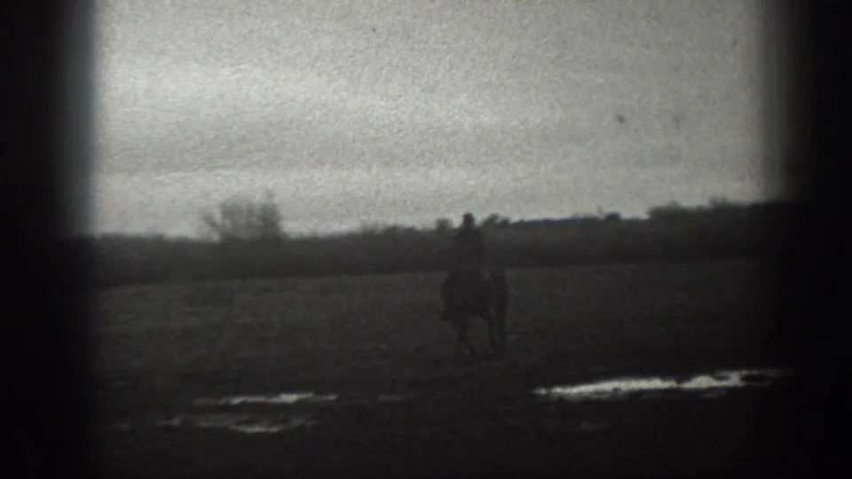 A man on horseback appears nearer in the frame, crossing the muddy Montana field in soft black-and-white 8mm footage.