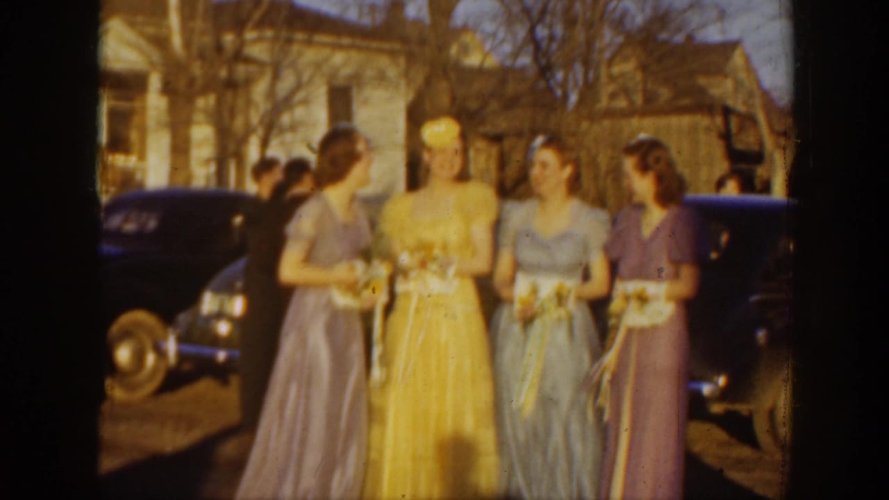 Four women in pastel gowns hold bouquets beside parked dark cars and a sunlit building.