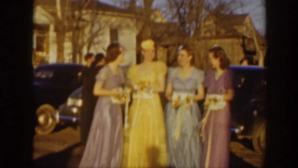 In 1939 Montana home-movie footage, four women in formal pastel gowns stand with bouquets near dark cars and a residential-looking building.