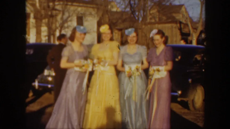 Four women in pastel formal dresses hold bouquets while standing outdoors before rounded 1930s automobiles in the Montana home-movie scene.