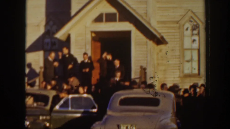 1939 Montana wedding guests stand near a wood-sided church entrance while rounded classic cars fill the foreground.