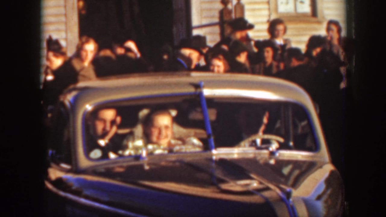 Newlyweds and an older driver sit in a vintage car as wedding guests gather behind them.