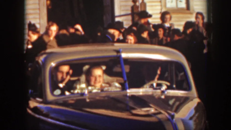 In the supplied 1939 Montana wedding footage, the bride, groom, and driver sit in a chrome-trimmed car before a crowd of guests.