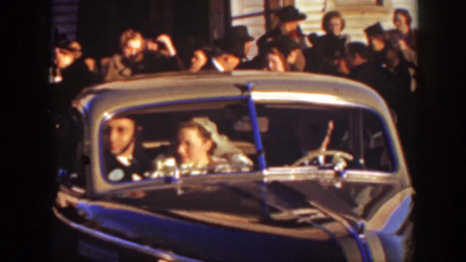 The bride and groom appear in the front seat of a prewar-style car during the Montana wedding sendoff, with guests gathered outside.