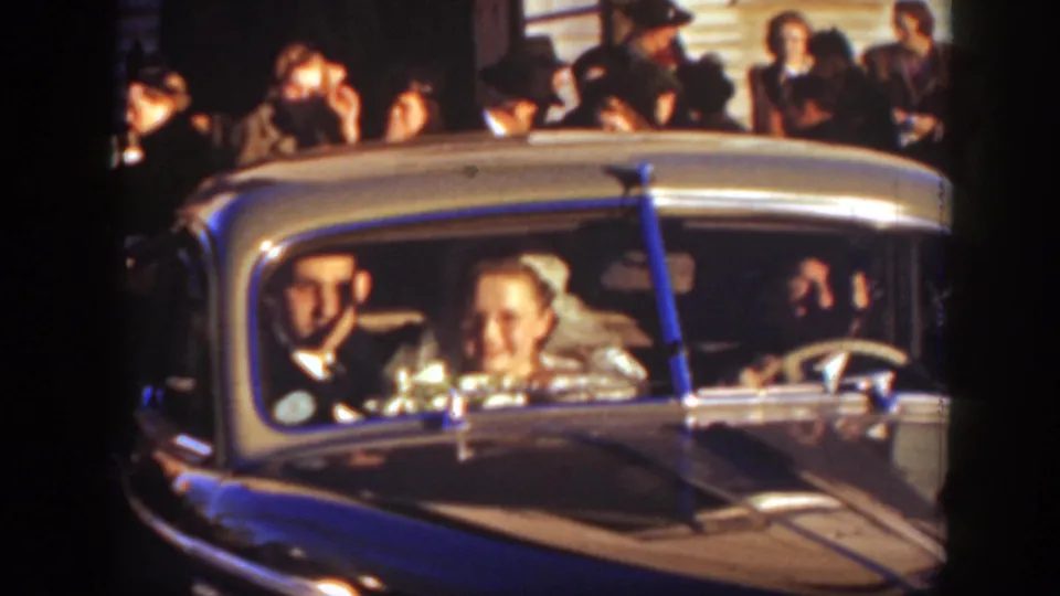 The bride smiles beside the groom in the vintage car, with a wedding crowd visible behind them in the 1939 Montana home movie.
