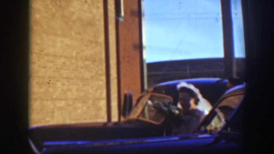 1939 Montana wedding footage shows the bride visible through or beside a car near brick buildings and a shadowed doorway.