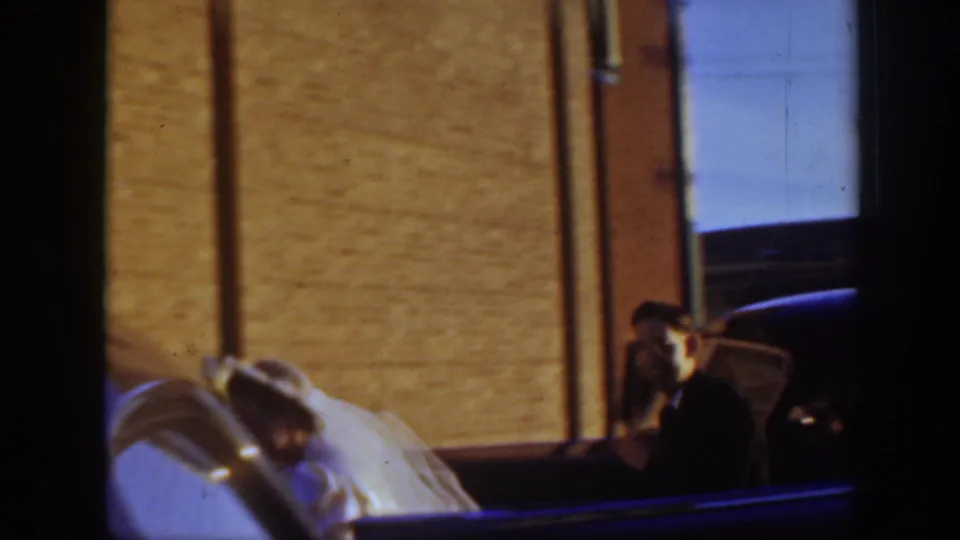The bride in a white gown and veil leans or steps beside the car while a suited man is partly visible near brick walls.