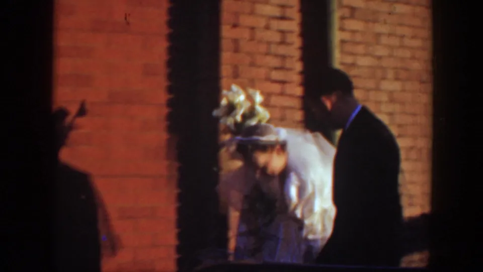 The bride in a white gown and the groom in a dark suit walk together beside brick walls in the 1939 Montana home movie.