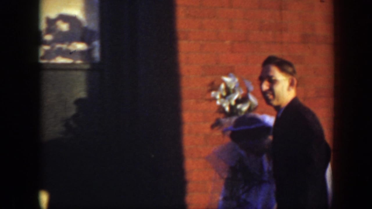 In 1939 Montana home-movie footage, a bride in a white gown and veil stands near a red brick church wall while a man in a dark suit faces the camera.