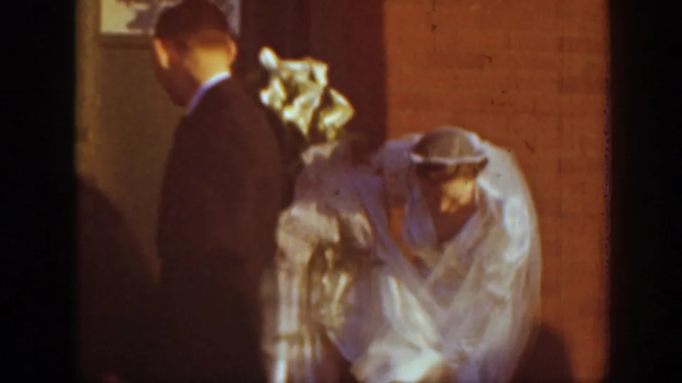 A suited man stands close to a veiled bride beside a red brick church entrance in the Montana wedding home movie.