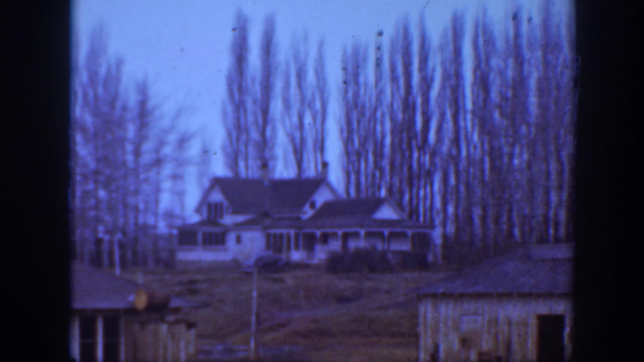 1939 Montana 8mm frame showing a white hillside farmhouse with a porch, bare trees, and a small foreground outbuilding.