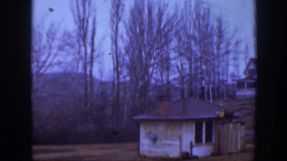 1939 Montana 8mm view with a small outbuilding in front and a distant hillside house partly obscured by leafless trees.