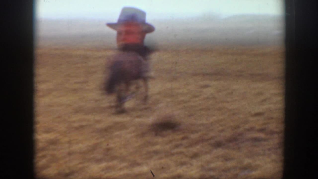 Rider on horseback moves away across open grassland, seen in soft focus through the film frame.