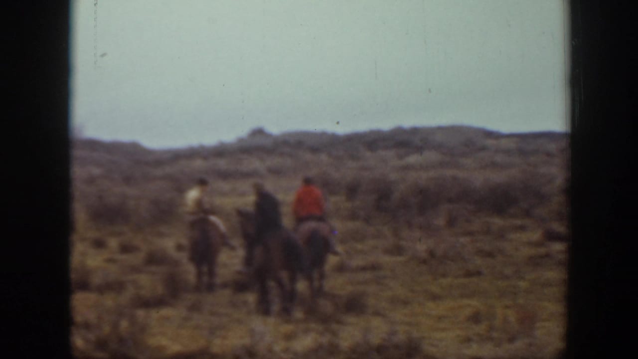 In 1939 Montana home-movie footage, three riders on horseback travel away across sandy open ground with low brush and distant hills.